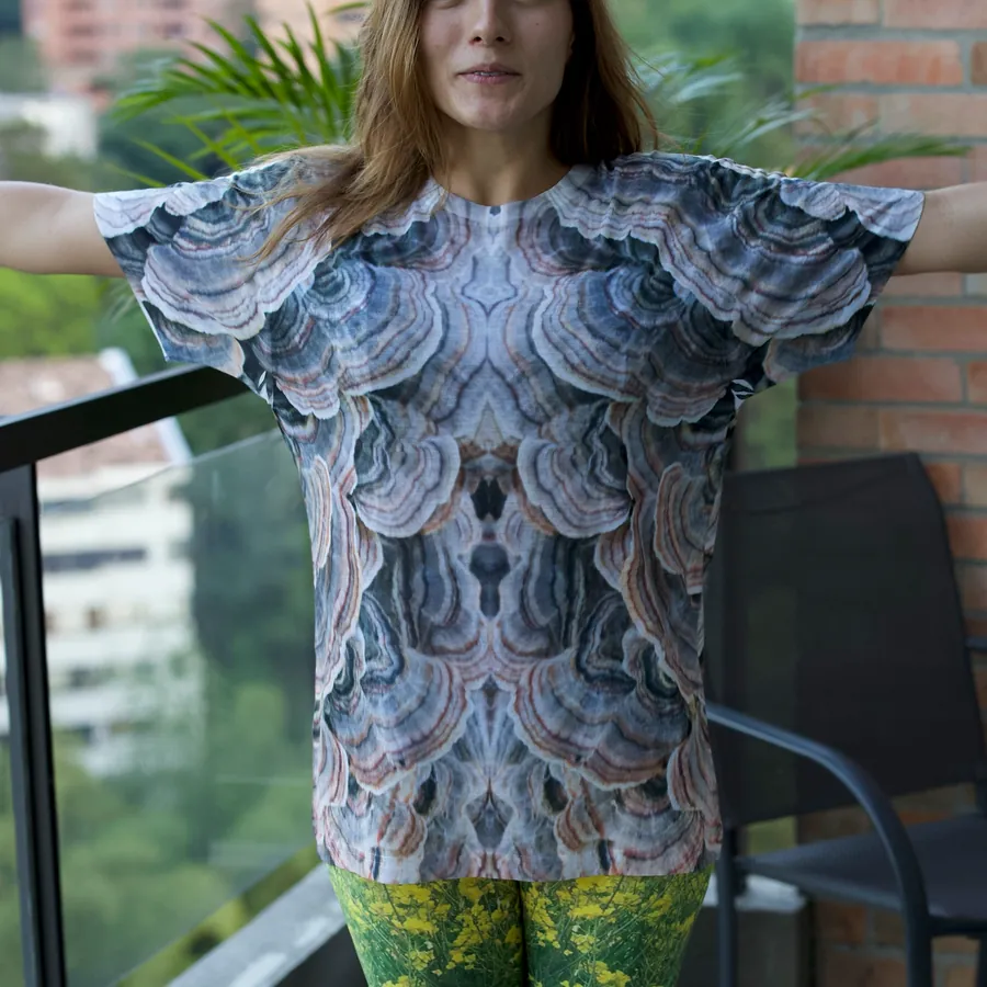 A model on a Medellín balcony wearing the Turkey Tail tee — an all-over print of bracket-fungus rings in grey, rust and cream — with green wildflower leggings.