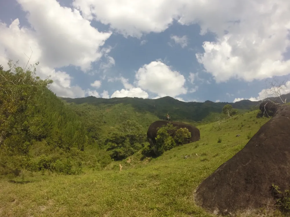 Green hills and granite boulders under high cloud in the Colombian countryside, with a figure standing on the largest boulder.